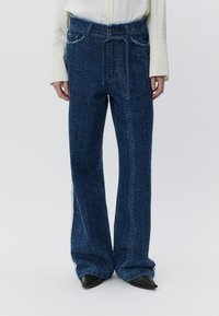 Niebieskie spodnie o szerokich nogawkach z fakturą, z paskiem ściągającym w talii, rozcięciami po bokach i widocznymi detalami szwów, połączone z jasną koszulą.