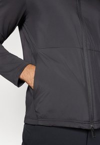 Jaqueta isolada preta com fecho de correr na frente, mangas longas e um bolso lateral. Tecido suave com um leve brilho e costura em contraste.