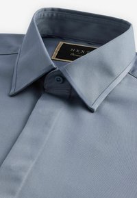 Camisa de manga corta azul claro con cuello puntiagudo, botón frontal único y una textura de tela suave y mate. Etiqueta de la marca visible dentro del cuello.