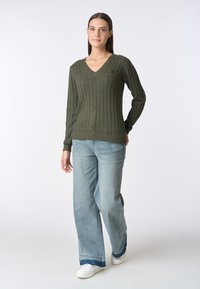 Maglione verde oliva a lavorazione cable knit con scollo a V e orlo a costine abbinato a jeans a gamba larga blu chiaro. Sneakers bianche con dettagli blu.