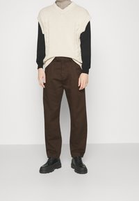 Person klädd i en beige stickad väst över en svart långärmad tröja, bruna byxor och svarta klumpiga skor, stående mot en vit bakgrund.