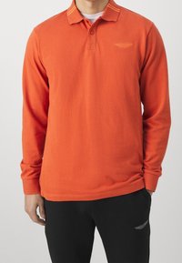 Pitkähihaine polo paita kirkasta oranssia puuvillaa, jossa on napitus, ribattu kaulus ja logon brodeeraus rinnassa. Yhdistetty mustiin housuihin.