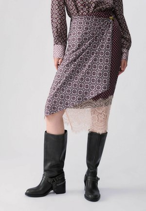 Donna che indossa una gonna a portafoglio fantasia con bordino in pizzo, abbinata a una camicetta a maniche lunghe coordinata e stivali neri in pelle al ginocchio.