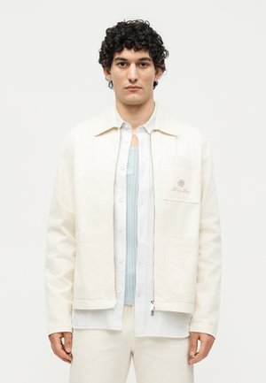 Mand med krøllet mørkt hår iført en cremefarvet jakke over hvid skjorte og lyseblå striktop, stående mod en ensfarvet off-white baggrund.