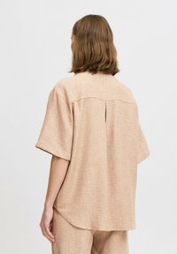 Košeľa s krátkym rukávom v svetlej taupe farbe, vyrobená z textúrovanej tkaniny. Má zadný yoke so subtílnou záhyb a zaoblený lem.