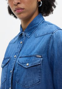 Camisa de ganga em um tom de azul médio, com colarinho pontudo, botões de pressão e dois bolsos no peito com aba e marca. Tecido texturizado.