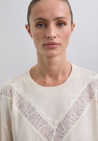 Junge Frau mit zurückgekämmtem Haar, die kleine Creolenohrringe und einen cremefarbenen Pullover mit einem V-förmigen Spitzen-Detail auf der Brust trägt.