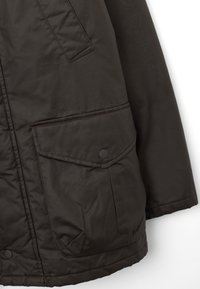 Giacca imbottita marrone scuro con finitura testurizzata, caratterizzata da due tasche frontali, chiusure a scatto e dettagli cuciti lungo le cuciture.