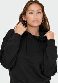 Felpa nera con cappuccio, in cotone, presenta un cappuccio con cordino, polsini a coste e una tasca a marsupio. Texture morbida con vestibilità rilassata.
