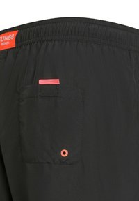 Pantaloni da nuoto neri con elastico in vita, dotati di una tasca posteriore con un'etichetta rosa e un piccolo dettaglio ad occhio arancione. Materiale leggero.