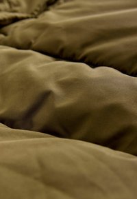 Detailní záběr na texturovanou, olivově zelenou polstrovanou látku s jemnými záhyby a mírnými vráskami, která naznačuje teplo a pohodlí.