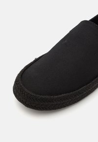 Svart slip-on sko med canvas overdel, rund tå og flettet jute detaljer langs sålen. Glatt tekstur uten synlige mønstre.