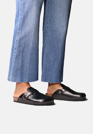Person iført lyseblå cropped jeans og sorte læder slip-on træsko med justerbare remme og korksåler på hvid baggrund.