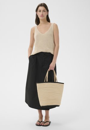 Femme portant un haut en maille sans manches beige, une jupe midi noire, des tongs noires, tenant un sac cabas tissé beige avec des bordures noires.
