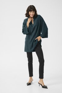 Tmavě tyrkysová tunika s výstřihem do V a tříčtvrtečními rukávy, v kombinaci s černými slim-fit kalhotami a černými mules na podpatku se dekorativními detaily.