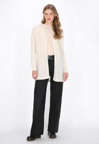 Lichtbeige cardigan met een open voorkant, lange mouwen en een zachte textuur, gecombineerd met hooggerokte zwarte jeans en zwarte schoenen.