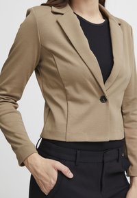 Kurt beige med enfaldig svart knapp, spetsiga slag och strukturerade ärmar, tillsammans med svarta högmidjade byxor.