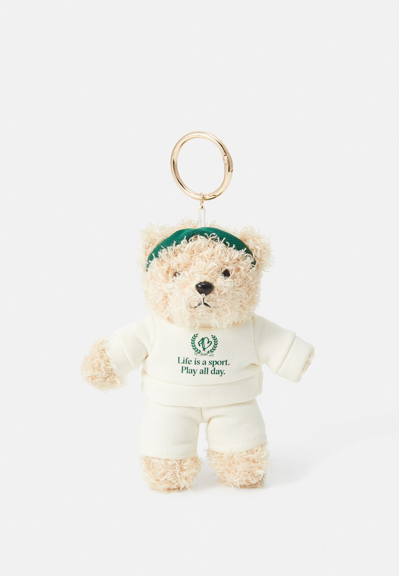 Soft plush bear keychain with light tan fur, wearing a white outfit and green headband. Features text "Life is a sport. Play all day."
