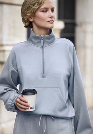 Hellgrauer Reißverschluss-Hoody mit hohem Kragen, elastischen Bündchen und Fronttasche. Hält einen Kaffeebecher mit schwarzem Deckel.