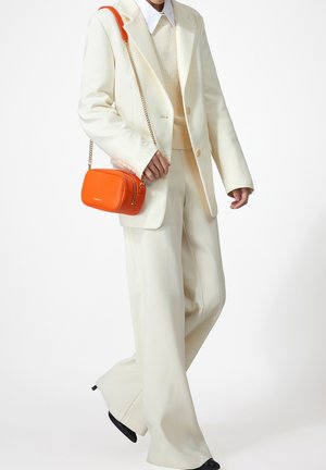 Persona con traje crema y suéter, sosteniendo una pequeña bolsa de cuerpo cruzado naranja con cadena dorada, de pie frente a un fondo blanco.