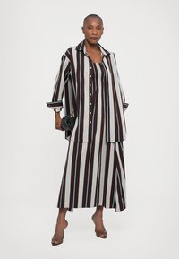 Striped maxi shirt dress in brown, white, and light blue. Features large collar, button front, and side slit. Paired with black handbag and heels.
