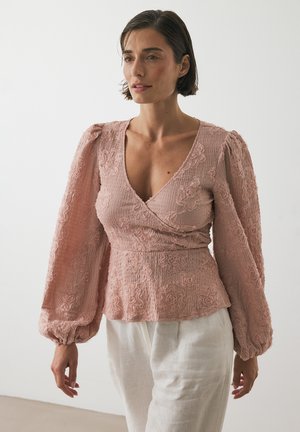 Femme portant un blouse enroulée texturée rose poussiéreux avec des manches longues bouffantes et un pantalon en lin blanc, se tenant devant un fond uni.
