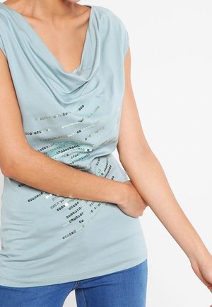 Person iført en lyseblå ærmeløs top med pailletter og blå jeans, arm bøjet med hånden hvilende nær taljen.