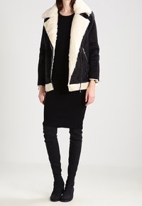 Czarny płaszcz z owczej wełny z kremowym futrzanym wykończeniem; ma szeroki kołnierz, zamek błyskawiczny z przodu oraz kieszenie po bokach. Noszony na czarnej sukience i wysokich kozaczkach.