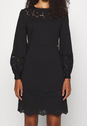 Vrouw in een zwarte jurk tot op de knie met kanten details op de halslijn, mouwen, taille en zoom, staand tegen een neutrale achtergrond.