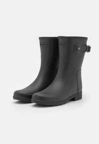 Botas de lluvia de caucho negro con una textura lisa, altura hasta la mitad de la pantorrilla, punta redonda y una correa lateral. Presenta un logo en relieve en la parte frontal.