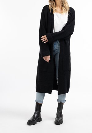 Personne portant un long cardigan noir, un t-shirt blanc, un jean bleu clair retroussé et des bottes noires épaisses, debout devant un fond blanc.