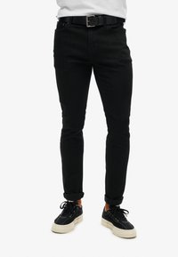 Pantalones de mezclilla negra con un corte ajustado, que presentan un diseño clásico de cinco bolsillos, detalles de costura y puños enrollados. Combinados con zapatillas negras.