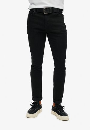 Pantalones de mezclilla negra con un corte ajustado, que presentan un diseño clásico de cinco bolsillos, detalles de costura y puños enrollados. Combinados con zapatillas negras.