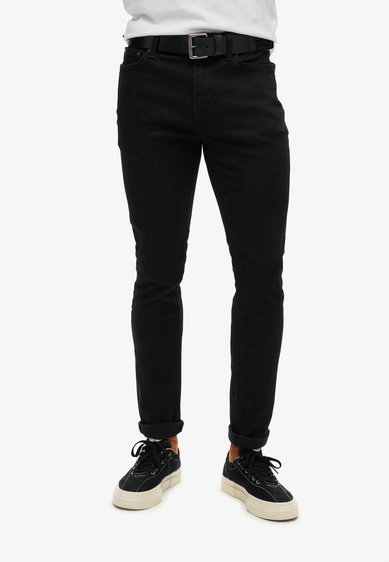 Pantalones de mezclilla negra con un corte ajustado, que presentan un diseño clásico de cinco bolsillos, detalles de costura y puños enrollados. Combinados con zapatillas negras.