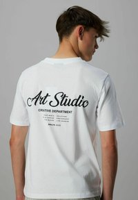 Jonge man die wegkijkt en een wit T-shirt draagt met zwarte tekst "Art Studio Creative Department" en kunstgerelateerde woorden op de achterkant.