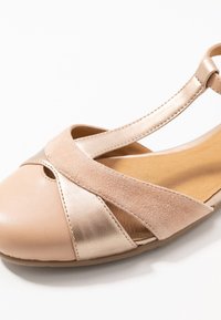 Sandal de plano abierto en beige y dorado metálico con correas entrecruzadas y correa en el tobillo sobre un fondo blanco.