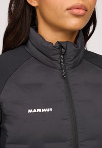 Svart vadderad jacka med hög krage, med en framdragkedja, loggodatum och textilstruktur. Släta detaljer på axlarna.