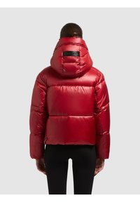 Henkilö, joka on pukeutunut kiiltävään punaiseen hupulliseen toppatakkiin, nähtynä takaa yksinkertaisen harmaan taustan edessä.