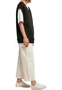 Czarna, oversizeowa bezrękawnikowa bluza z dekoltem w serek, zestawiona z warstwową białą koszulą pod spodem oraz luźnymi białymi spodniami, noszona z białymi trampkami.