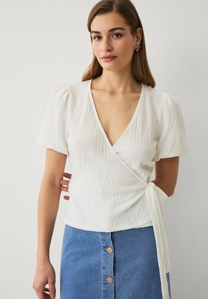 Vrouw draagt een witte, gestructureerde, korte mouw wikkelblouse met een zijstrik en een blauwe denimrok met knopen aan de voorkant, staand tegen een effen muur.
