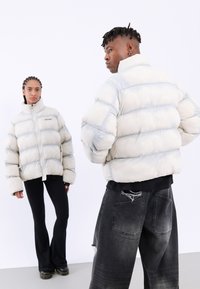 Ljust grå puffjacka med horisontella sektioner, hög krage och framdragkedja. Bärs med svarta utsvängda byxor som har en slitna denim-look.