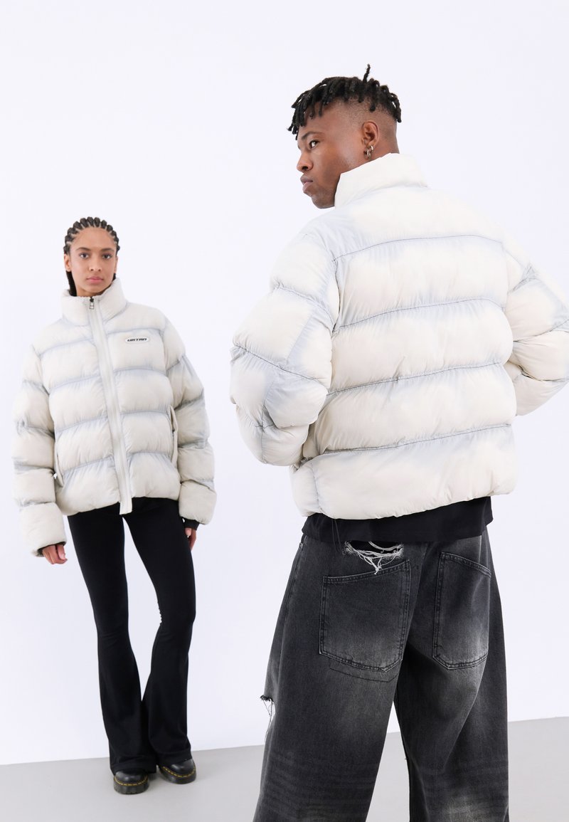 Ljust grå puffjacka med horisontella sektioner, hög krage och framdragkedja. Bärs med svarta utsvängda byxor som har en slitna denim-look.