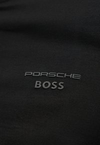 Μαύρο ύφασμα με ανάγλυφα μαύρα λογότυπα κειμένου "PORSCHE" και "BOSS" κεντραρισμένα στο υλικό.