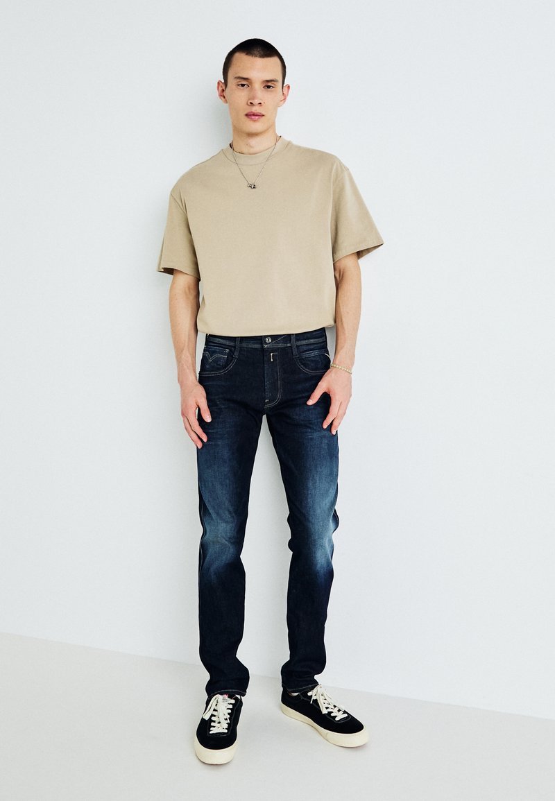 Modelo masculino em pé, vestindo uma t-shirt de manga curta bege, jeans escuros desbotados, ténis preto e branco, um colar e uma pulseira, em frente a uma parede branca.