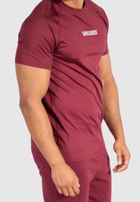 Röd atletisk t-shirt gjord av elastiskt tyg, med korta ärmar, rund halsringning och en kontrasterande vit logotyp på bröstet.