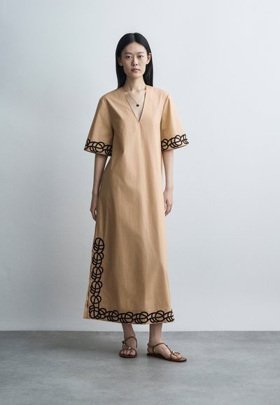 Femme debout portant une robe beige longue jusqu'aux chevilles avec une broderie ronde noire sur les manches et l'ourlet, assortie à des sandales marron à brides.