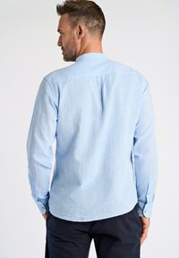 Jasnoniebieska koszula z długim rękawem o fakturowanym wykończeniu, z zapięciem na guziki przy mankietach oraz plisą z tyłu. Koszula ma zaokrąglony dół i kołnierzyk.