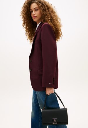 Kobieta z kręconymi włosami, ubrana w ciemnofioletową marynarkę i niebieskie dżinsy, trzymająca czarną torebkę za górny uchwyt przy boku.