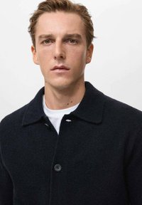 Abrigo de lana azul marino oscuro con cuello, cierre de botón único y textura sutil, llevado sobre una camisa blanca. Detalles de diseño simples.