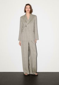 Lichte grijze pak met een gestructureerd ruitpatroon, voorzien van een dubbelzijdige blazer en wijde broekspijpen. Zwarte knoopdetails zichtbaar.